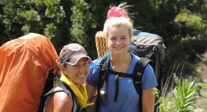 two people wearing backpacks smile while standing amid greenery 